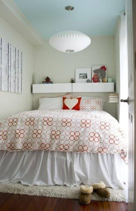 A bed squished between two walls with a red pillow on it with a white heart on the pillow and an oval sphere pendulant light.