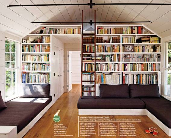 Books are stacked on white shelves in a room with at black sectional and a ladder.