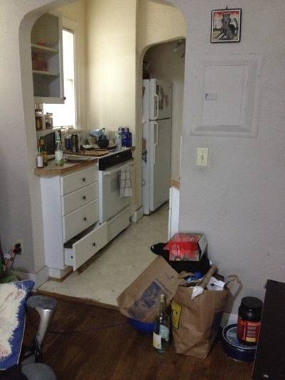 A couple of brown bags full of groceries next to a wall to a galley kitchen with lots of stuff on the counter over the stove.
