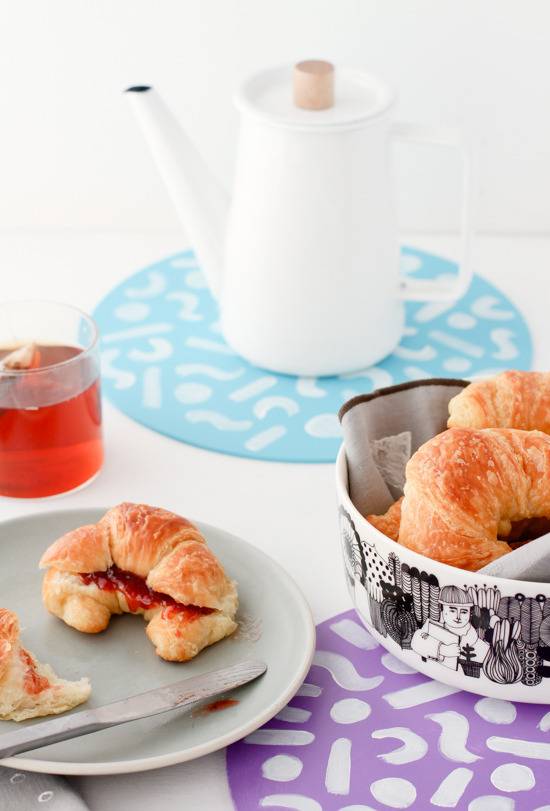 A croissant on a grey plante with a butter knife, a glass of orange liquid, a white coffee server on a blue and white round pad and a black and white bowl of croissants on a purple and white circle.