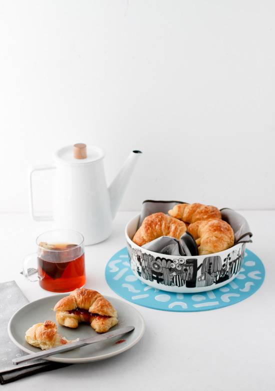 A plate with a croissant on it next to a knife, a glass of tea, a coffee woarmer and a black and white bowl full of croissants on a blue circle with white designs.
