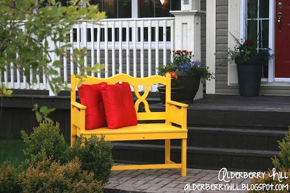 2 red throw pillows sit on top of a yellow bench in front of a white fence.