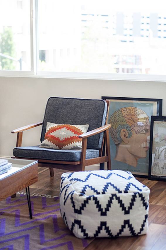 A white ottomon with black zig zags on it next to a wooden chair with grey cushions and a tan pillow with an orange diamond on it next to a piece of art with a mans head on a blue background.