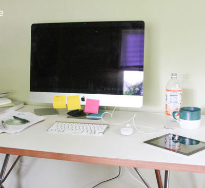 A computer monitor, keboard, a mouse, a bottle of water and a green and white mug on a white table.