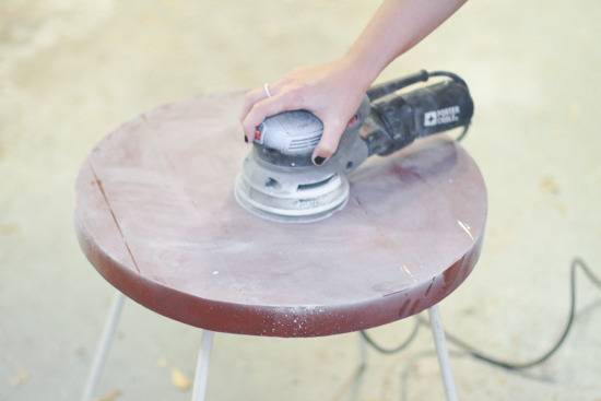 A hand using a round electric sander on the top of a round wooden stool.