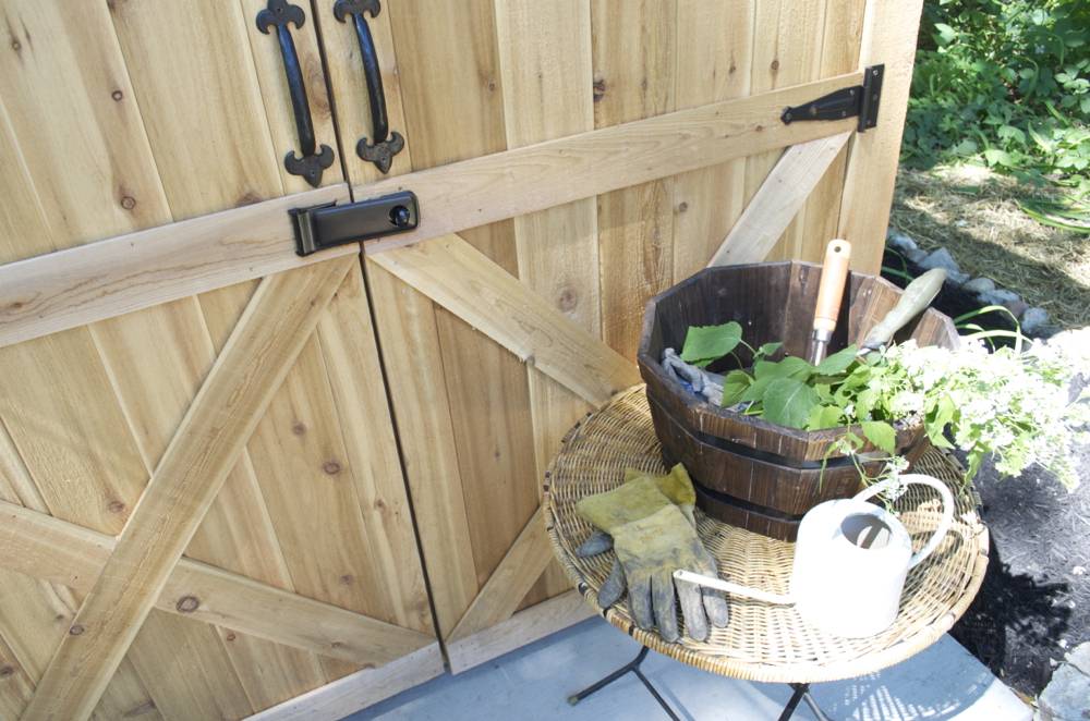 A wooden gate with a round wicker table in front of it holding a pair of yellow gardening gloves, a white watering can and a brown planter with gardening tools and a white flowering plant in it.