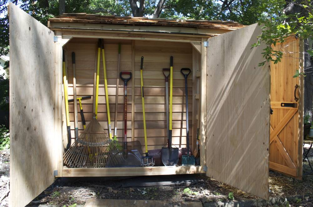An outdoor shed has hanging yellow garden tools.