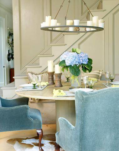 A light colored dining table with light blue arm chairs and blue hydrangeas on the table under a circular tea candle chandelier.