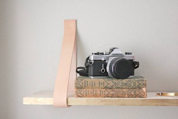 An antique camera on top of two books laying flat on a wooden shelf held up by a loop of light colored leather.