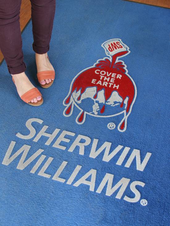 A woman stands next to an image of a paint can on a blue floor with Sherwin-Williams written on it.