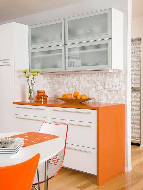 A kitchen has orange furniture and white walls.