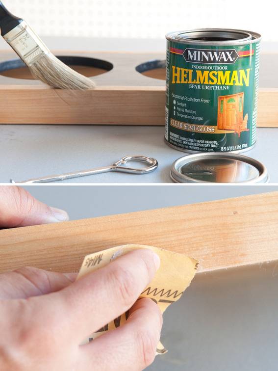 A hand sanding a piece of wood, a green can of wood stain and a paint brush painting a piece of wood with two large holes in it.