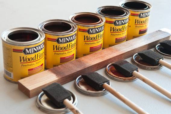 Five cans of wood stain lined up behind a length of wood and their tops holding black painting sponges.