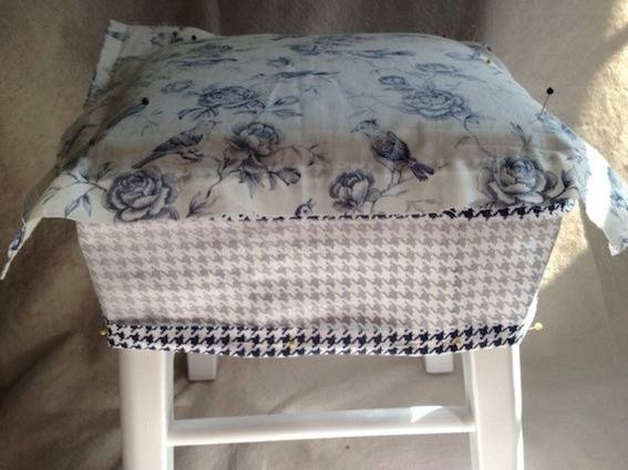 A blue and white floral fabric is on top of a stool that is covered with a blue and white hounds tooth fabric, and White painted legs.