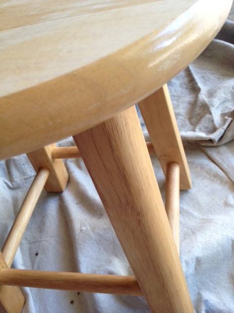 Edge of a stool made of light-colored wood, with a round seat and four legs.