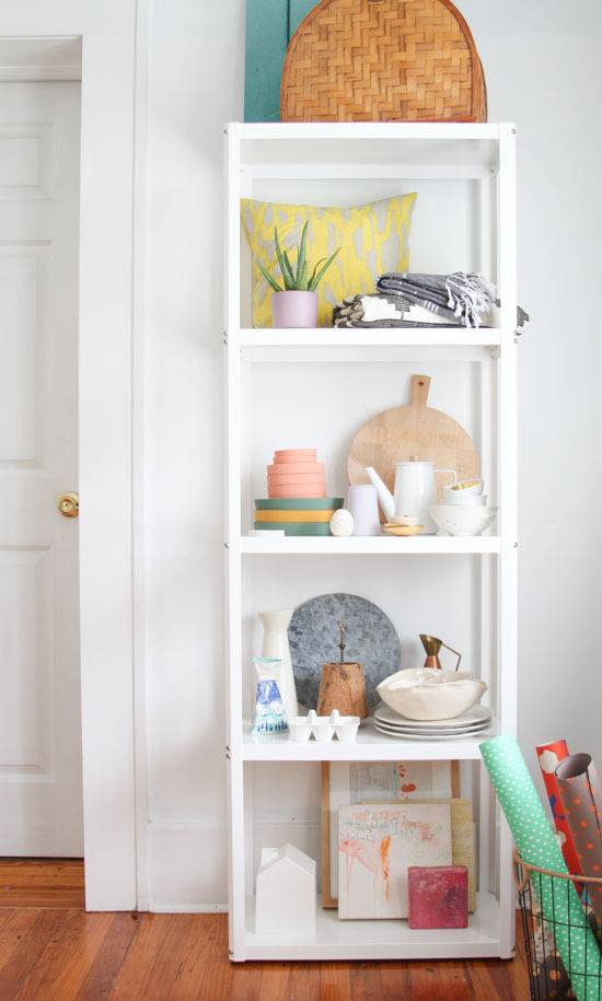 A four tiered shelf with a wicker basket on top and a yellow pillow on the top shelf.