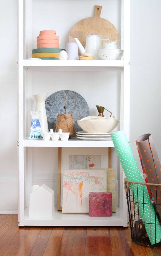 Knick knacks are arranged neatly on a white bookshelf next to a basket with rolled-up yoga mats.