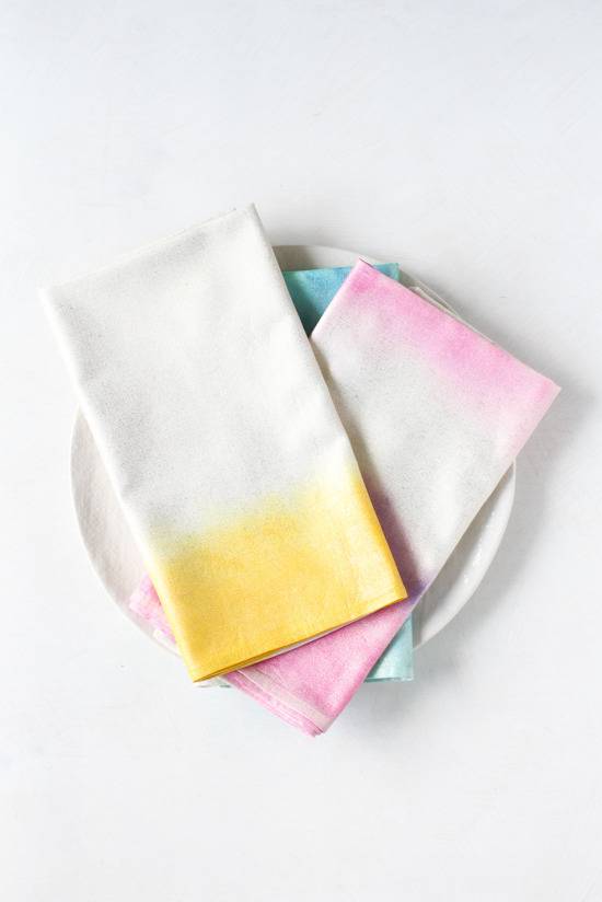 Three folded tye die napkins, one white and yellow, one pink and white and one blue and white on a white plate.