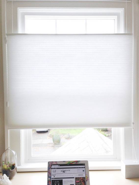 A laptop or tablet sts on a desk under a window with a white shade sitting in the center of the window.