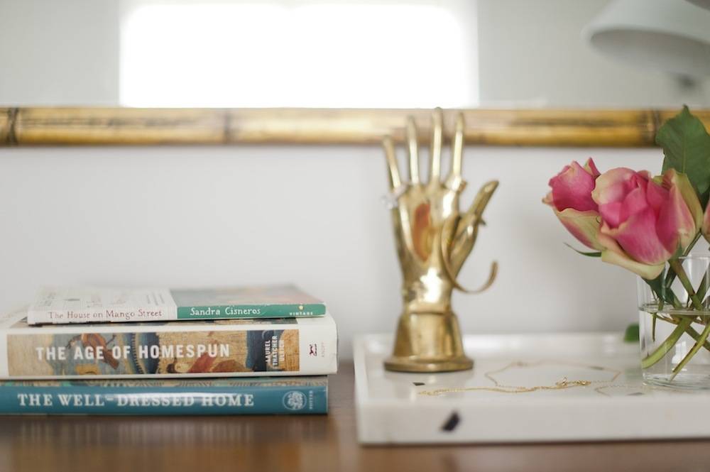 A golden hand with a gold bracelet hung on the thumb is in between two pink roses and a stack of three books.