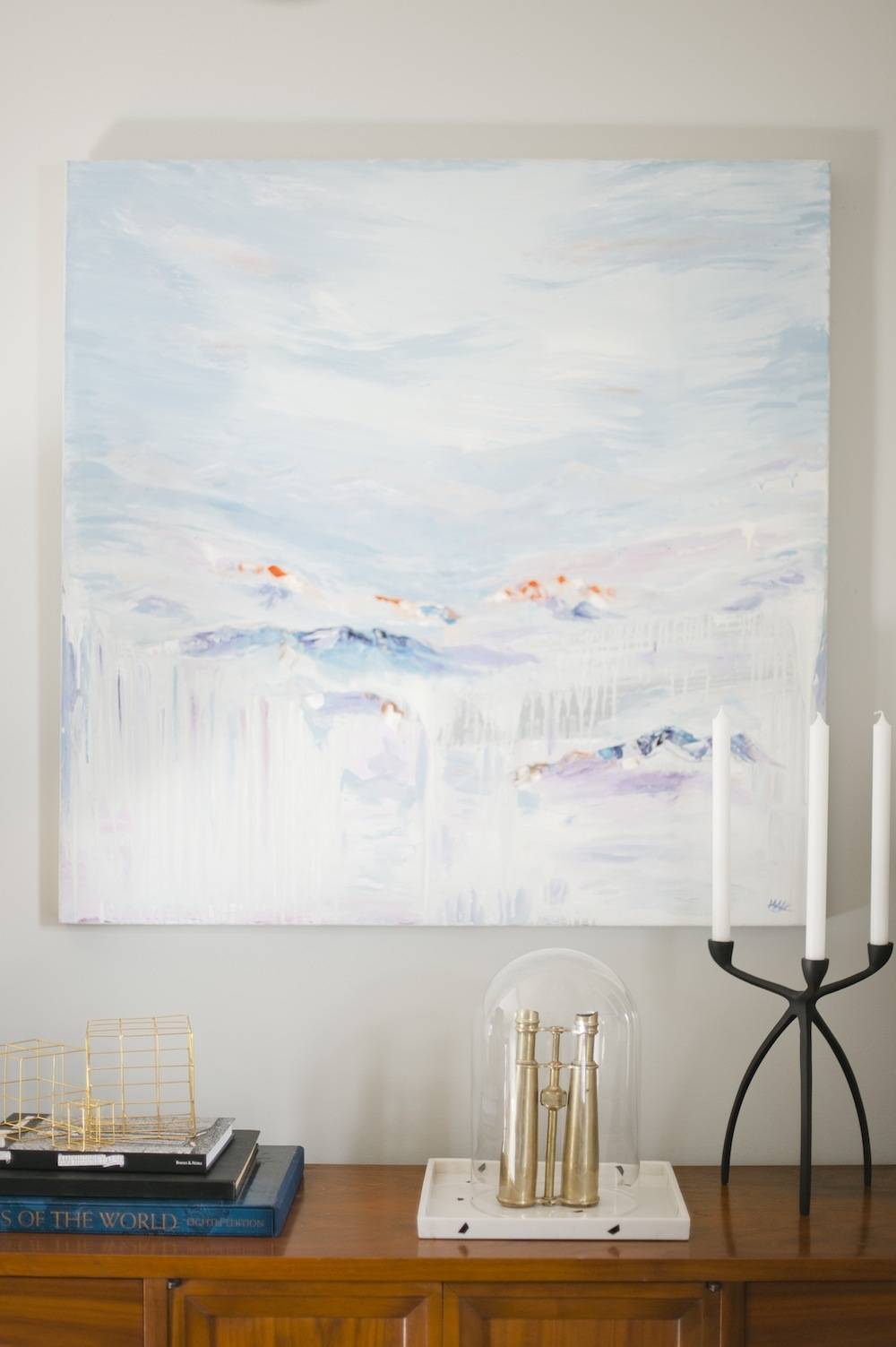 Pastel, watercolor-look artwork on a white wall above candlesticks and other items on a wood furniture piece.