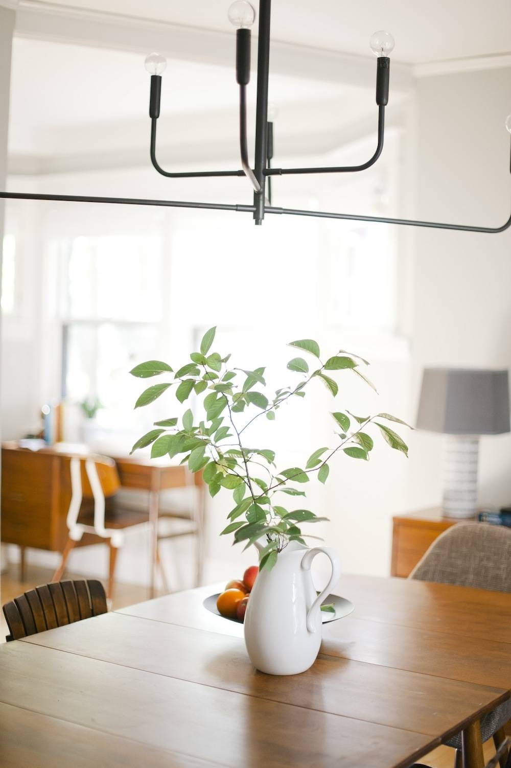 Room featuring dining table with foliage in a pitcher, under a modern four-pronged light fixture.