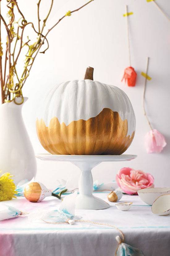 A pumpkin on a cake plate partially painted white, a white vase with yellow branches and pink and orange roses hanging upside down on the wall.