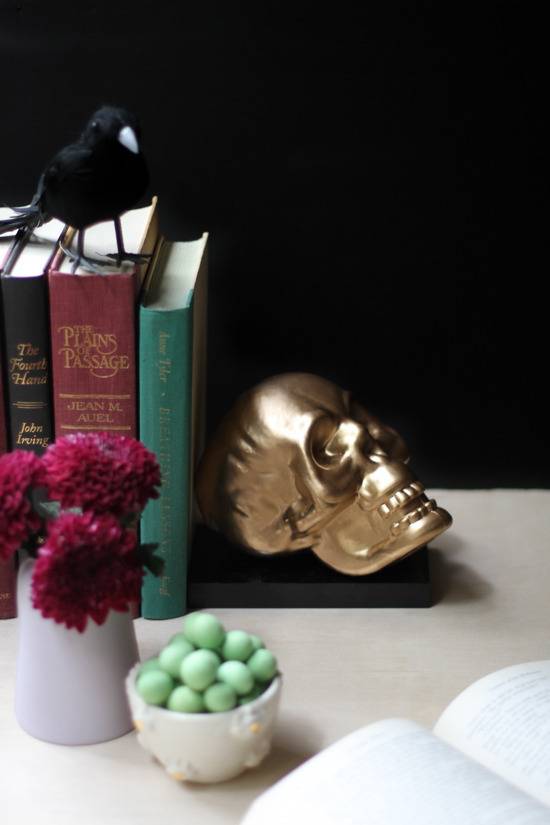 A gold skull bookend secures a red, green, and black book on a shelf, next to green mints, and red flowers.