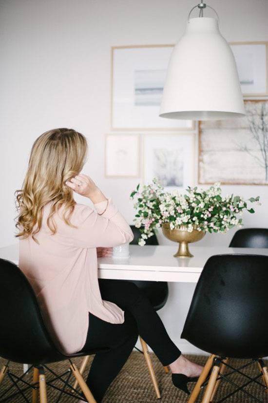 A blonde woman in a pink shirt and black pants is sitting in a black chair at at white table with three other black chairs, a white and green floral arrangement in a bronze cup on the table.