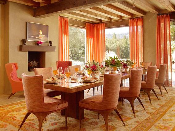 A long set dining table, with rounded orange chair and orange curtains on the tall windows and a rug with orange accents.