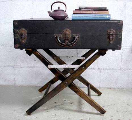 An antique suitcase table with a copper teapot on top as well as a stack of books laying sideways.