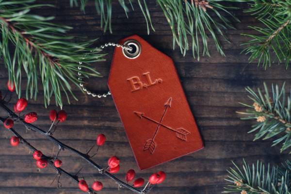 A red leather tag with the initials B.L. on it with crossed arrows in surround by pine twigs and red berries on a branch.