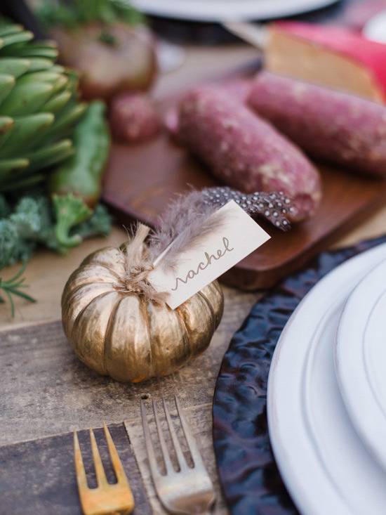 A small gold pumpkin with a name label on a table with two salami rolls and white plates on a blue placemat.