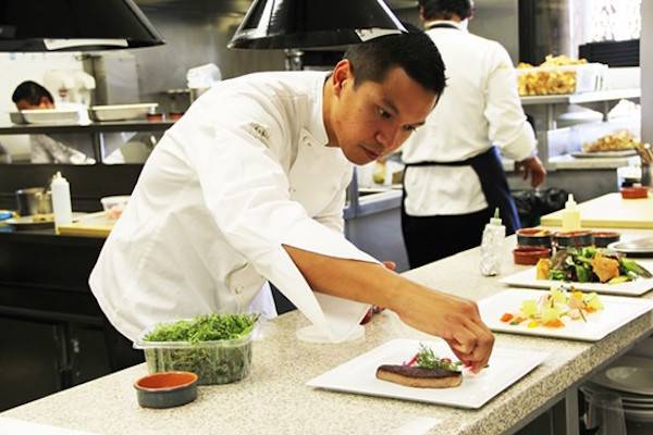 Chef bending over counter and carefully arranging food on a tray in a kitchen.