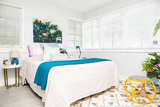 A bedroom with a yellow stool, a white bed with a blue throw, and a picture of a green tree above the bed.