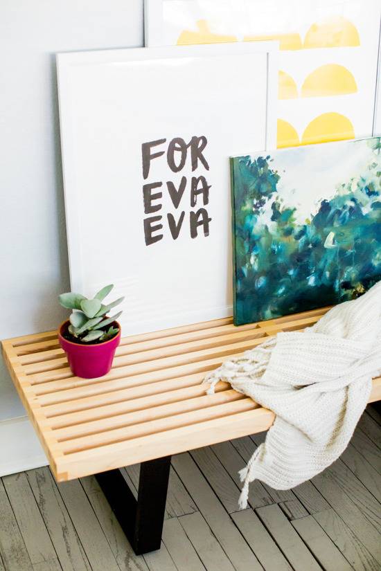 Three pictures, a small potted plant, and a throw blanket are on top of a wooden bench.