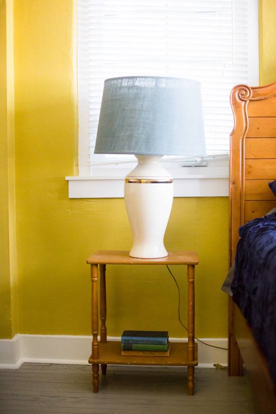 A white lamp sits on a wooden table in a yellow bedroom.