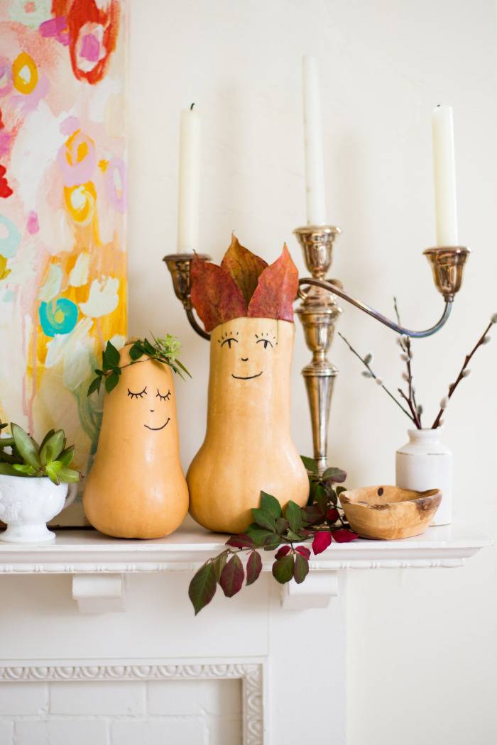 Two squash have smiley faces drawn on them and decorated as part of a festive arrangement on a fireplace mantel.