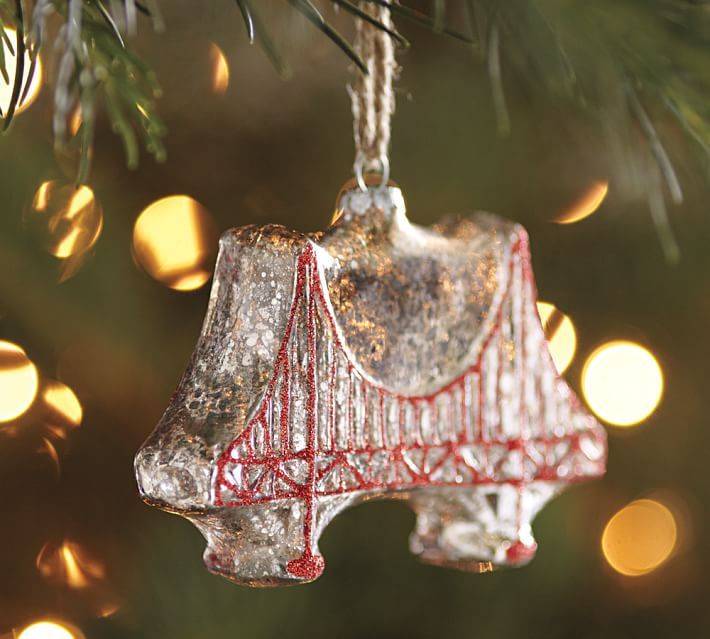Ornament in the shape of a suspension bridge, with red details, hanging from a holiday tree.