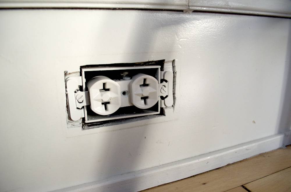 An electrical outlet on a white wall are uncovered.