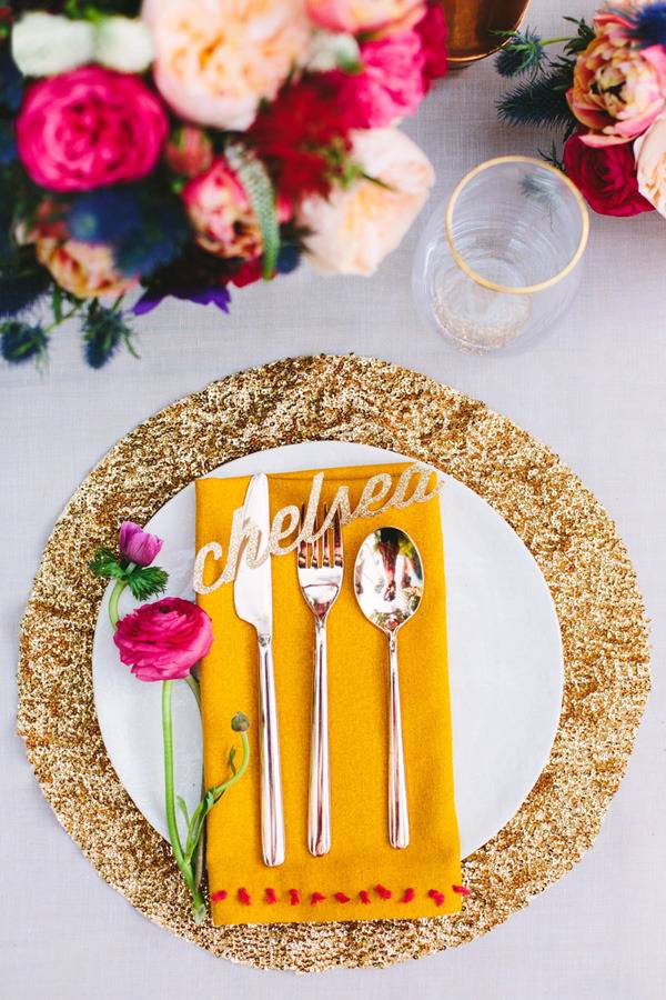A knife, fork and spoon sit on an orange folded napkin on a white plate on a golden round placemat next to a boquet of pink and peach carnations.