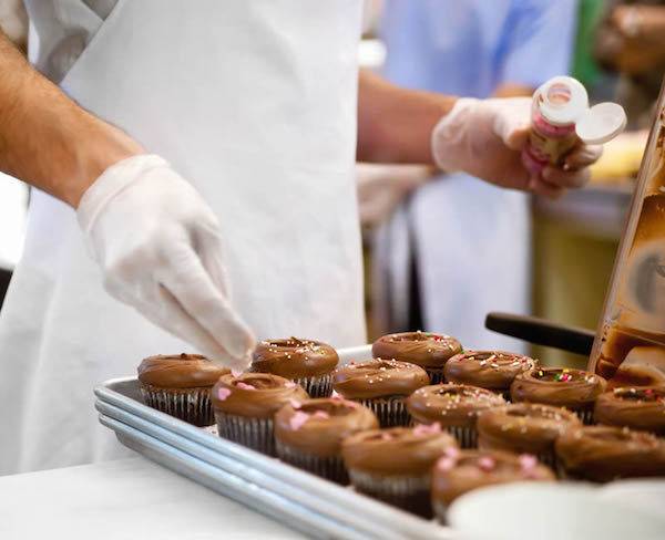 Person with gloved hands standing in front of a tray of chocolate cupcakes holding a bottle of edible decorations.