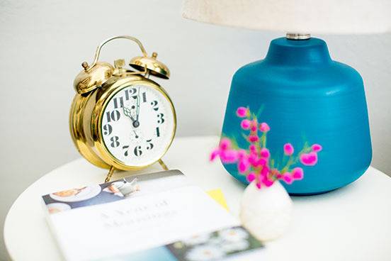 An alarm clock sits on a white table near a blue and white lamp.