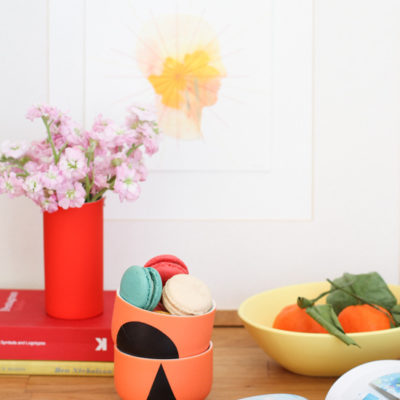 Red color flower vase with pink flowers, some books, orange fruits in a bowl, and some cookies at the top of the wooden table.