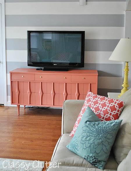A pink table with a television on it in a living room with a grey couch with one blue and one pink pillow on it.