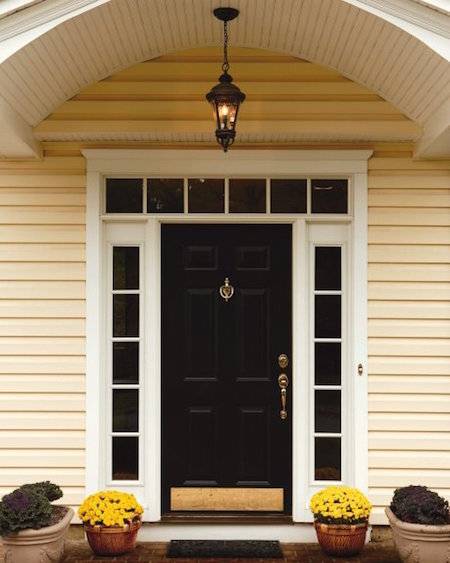 The front door to a cream yellow house is black, with pots of flowers on either side.