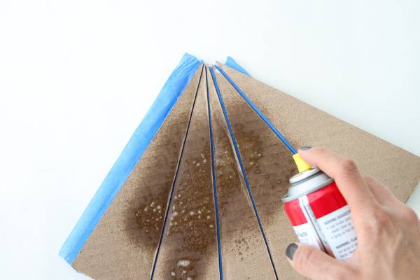 A lady uses a spray bottle to spray something onto cardboard with blue painter's tape on it.