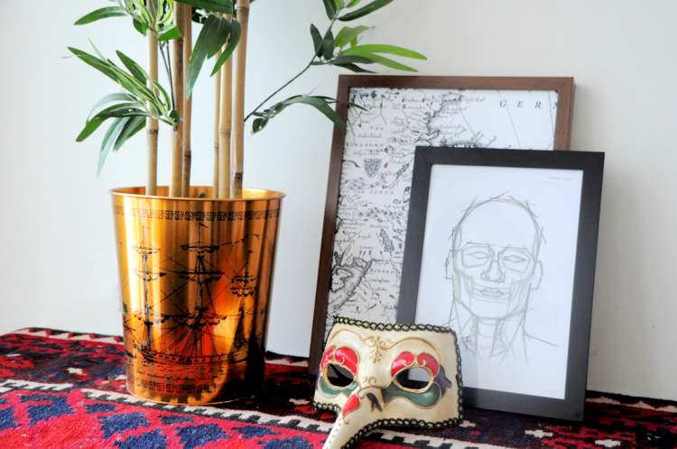 A potted plant, two framed drawings and a mask sitting on a desk.