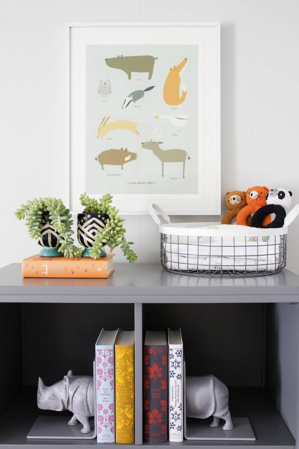 A gray shelf has books & plants on it under a picture frame.