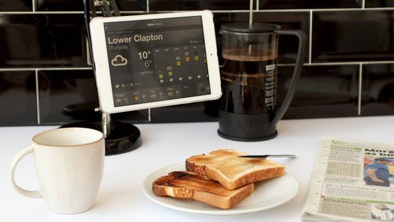 Toasted bread pieces in a white plate, coffee mug, news paper, coffee maker, and a tablet hanging to a stand are at kitchen counter top.